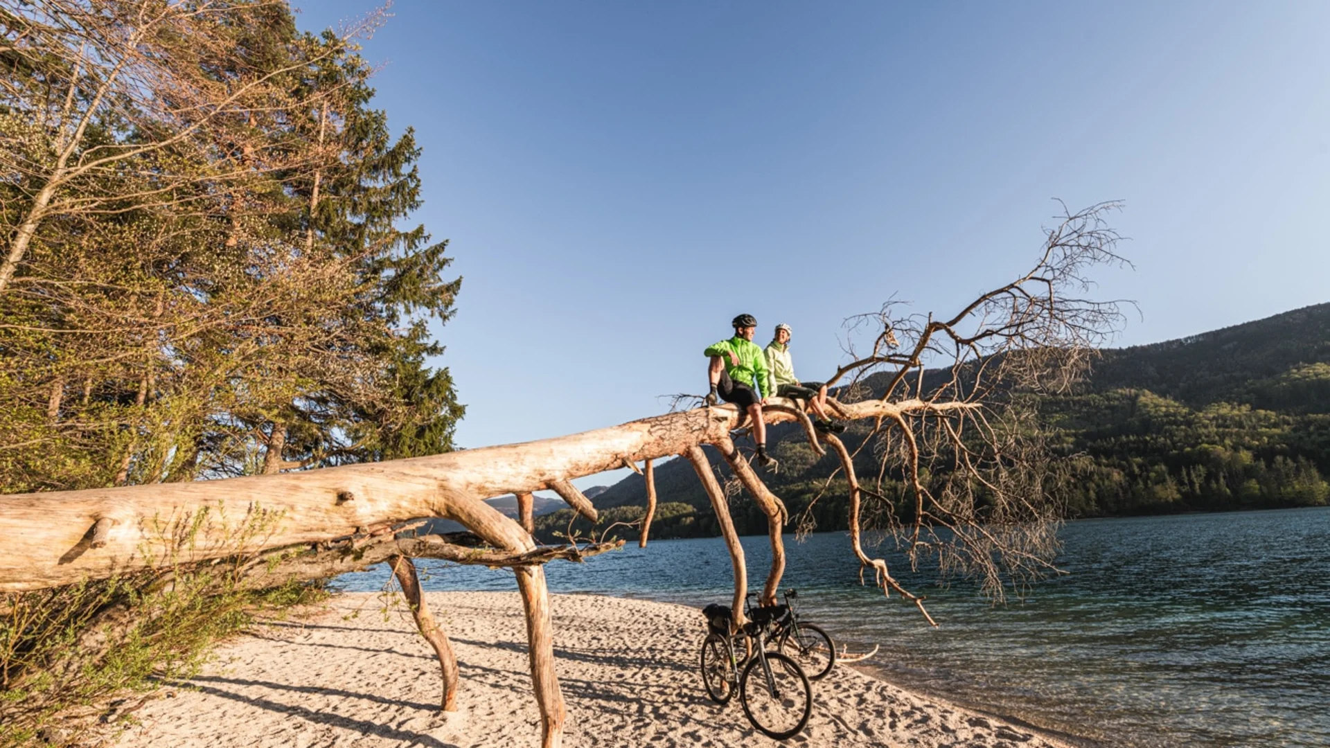 Two cyclists sitting on fallen tree by lake with mountains