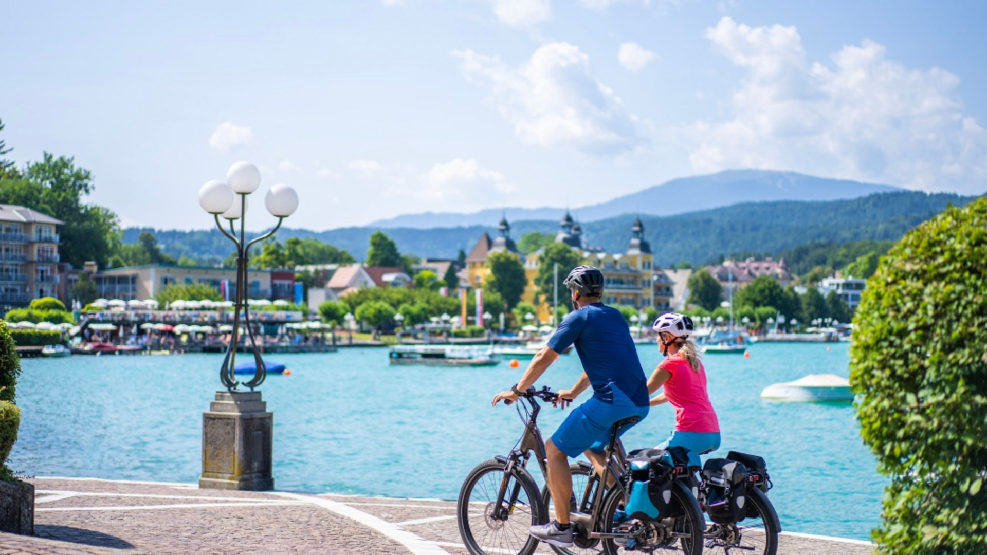 Two cyclists by the lakeside with mountains and buildings in the background