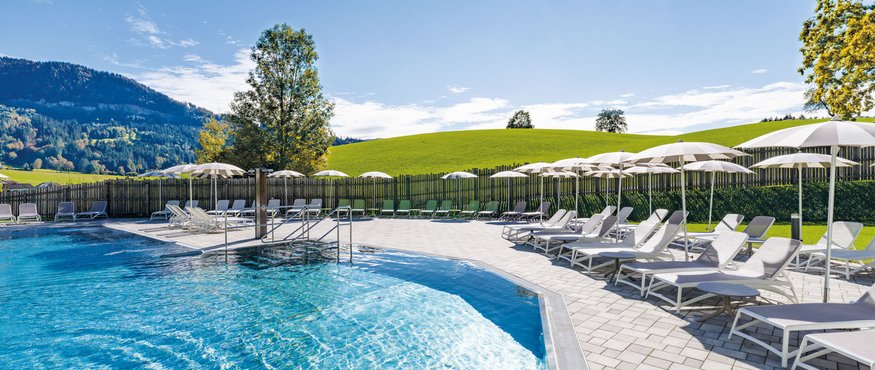 Empty pool with lounge chairs and umbrellas by green hills under sunny sky