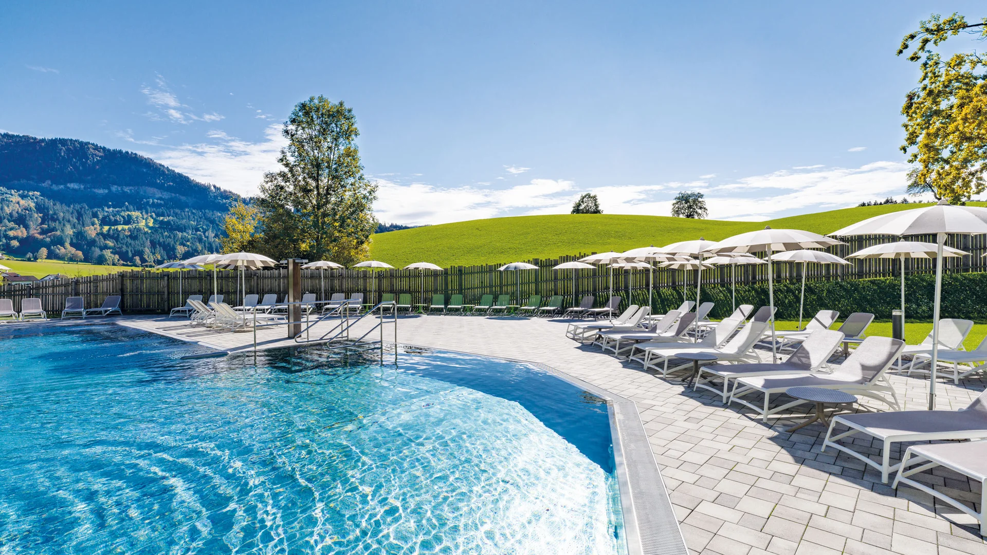 Leerer Pool mit Liegestühlen und Sonnenschirmen vor grünen Hügeln bei Sonnenschein