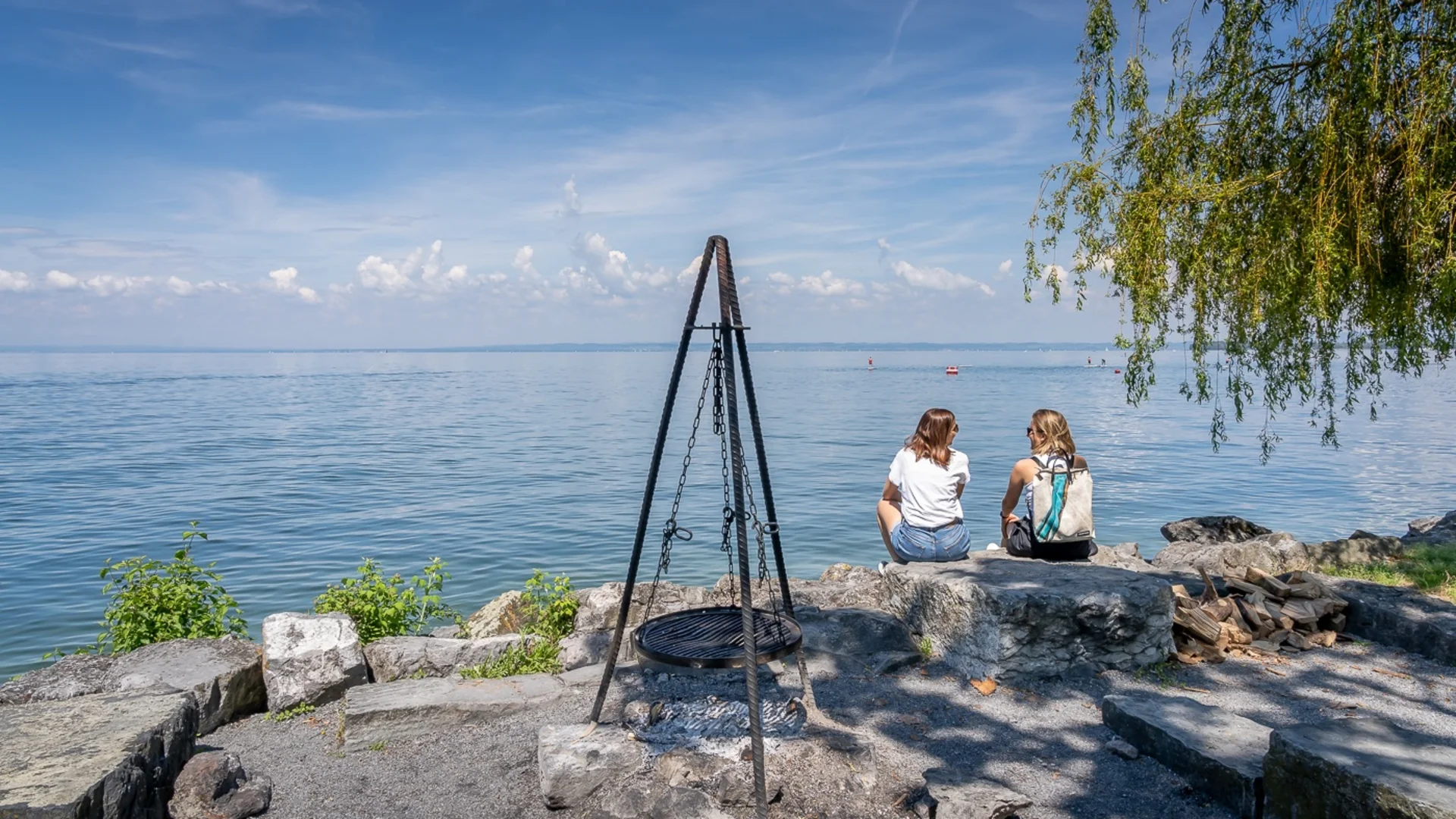 Zwei Frauen sitzen auf Steinen am See mit Grillrost und Holzstapel