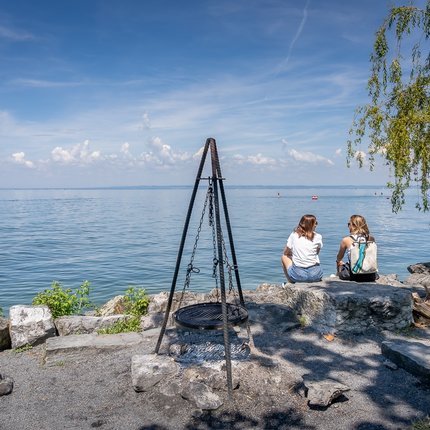 Two women sitting on rocks by the lake with grill and stacked firewood