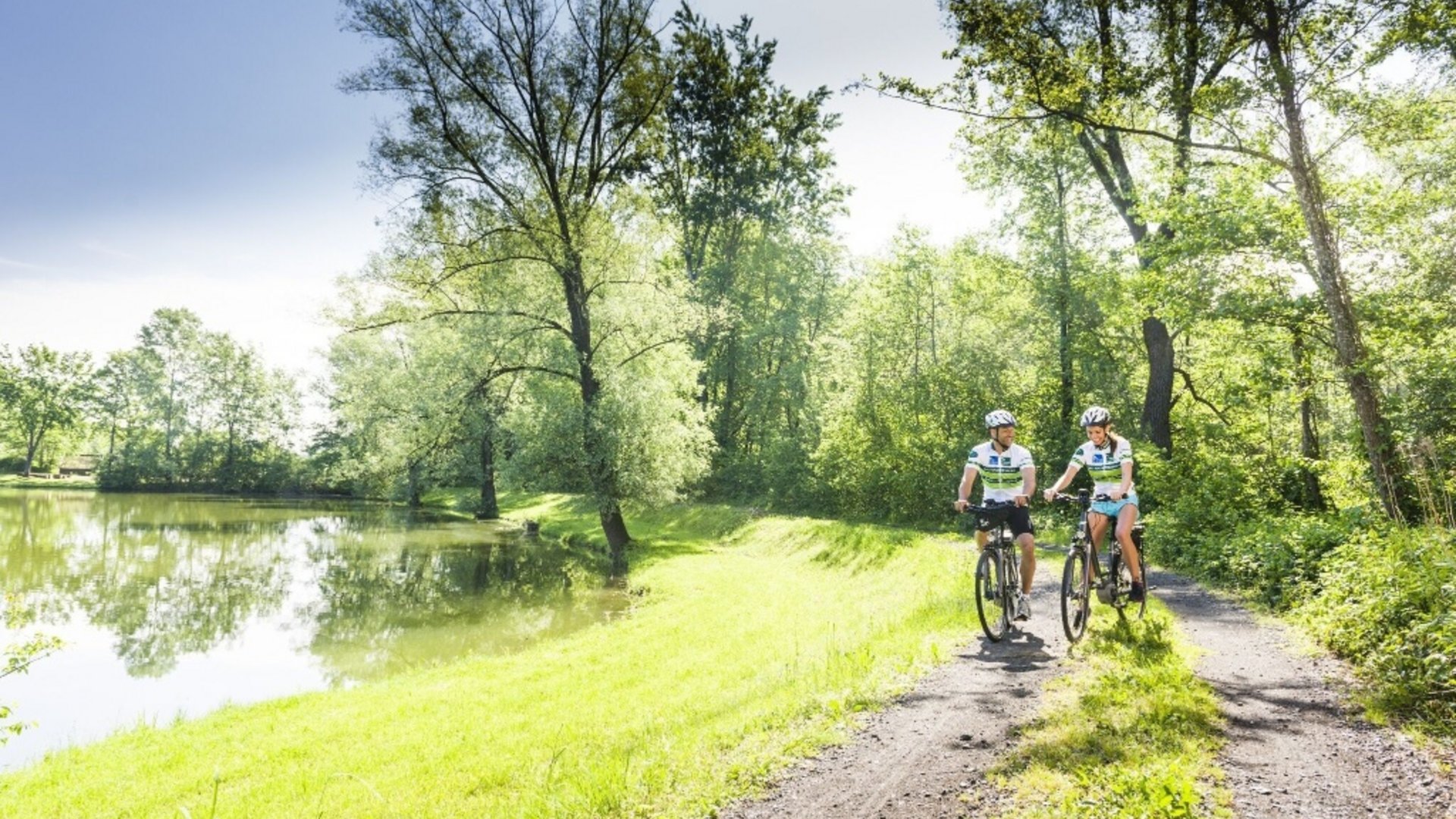 Two cyclists on forest path beside river on sunny day