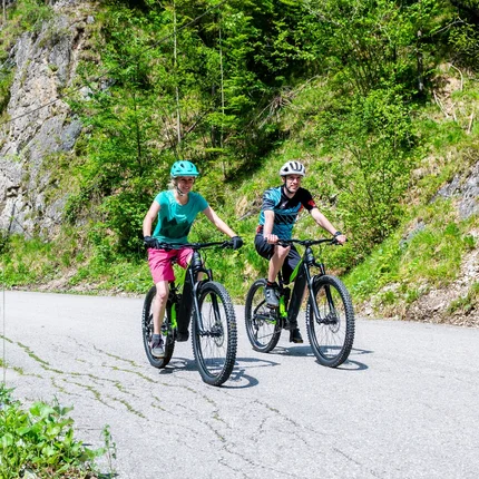 Twee mensen fietsen op mountainbikes over een bergweg langs groene rotsen