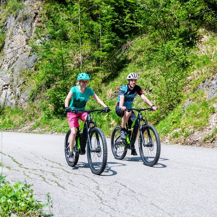 Two people riding mountain bikes on a mountain road with green rocky surroundings
