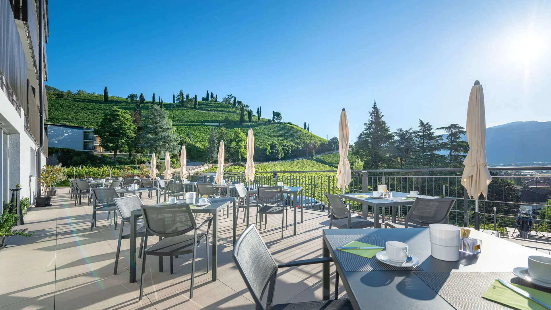 Sonnige Terrasse mit Tischen und Stühlen vor einem grünen Hügel
