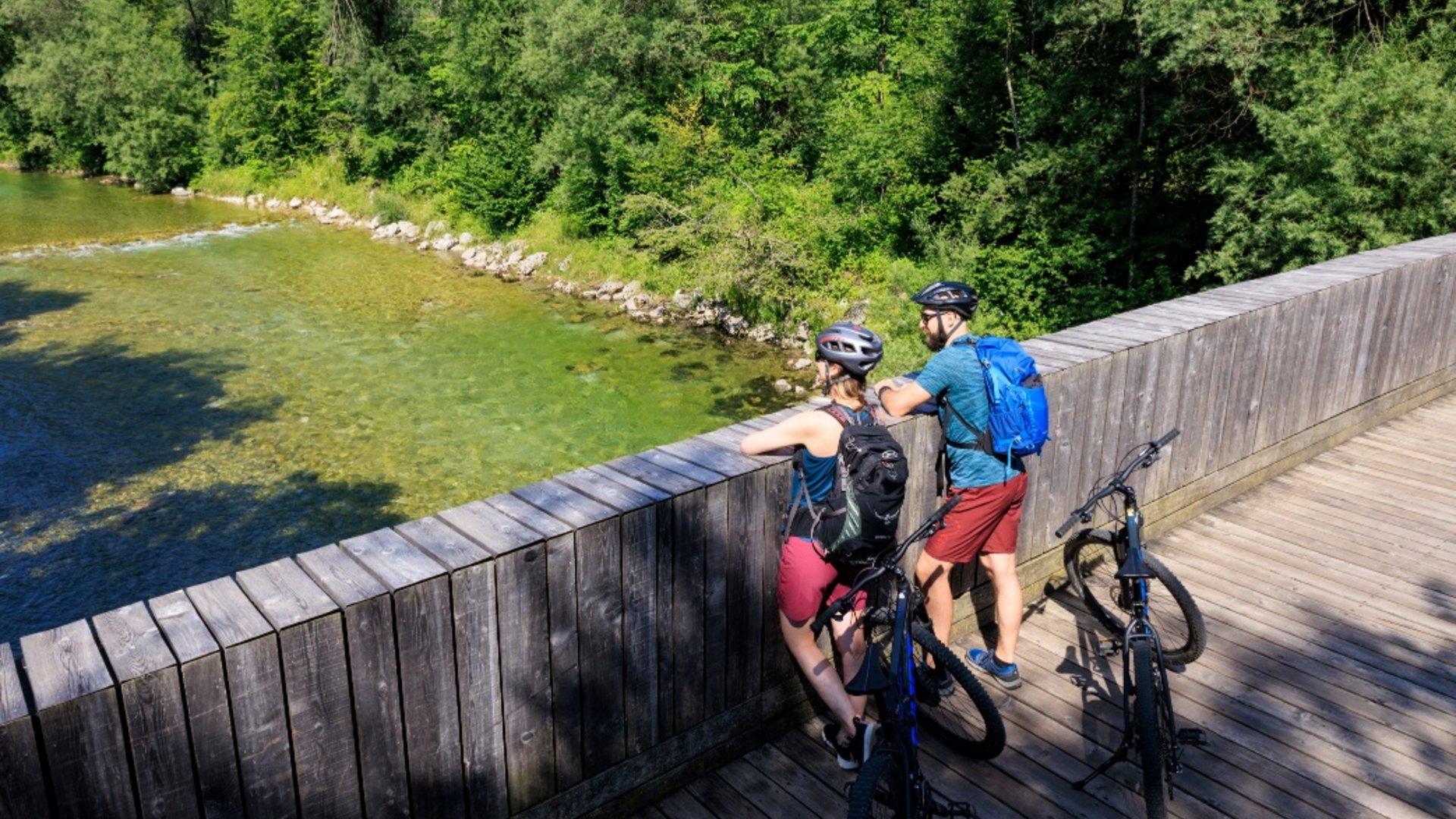 Two cyclists with helmets and backpacks on a wooden bridge over clear water