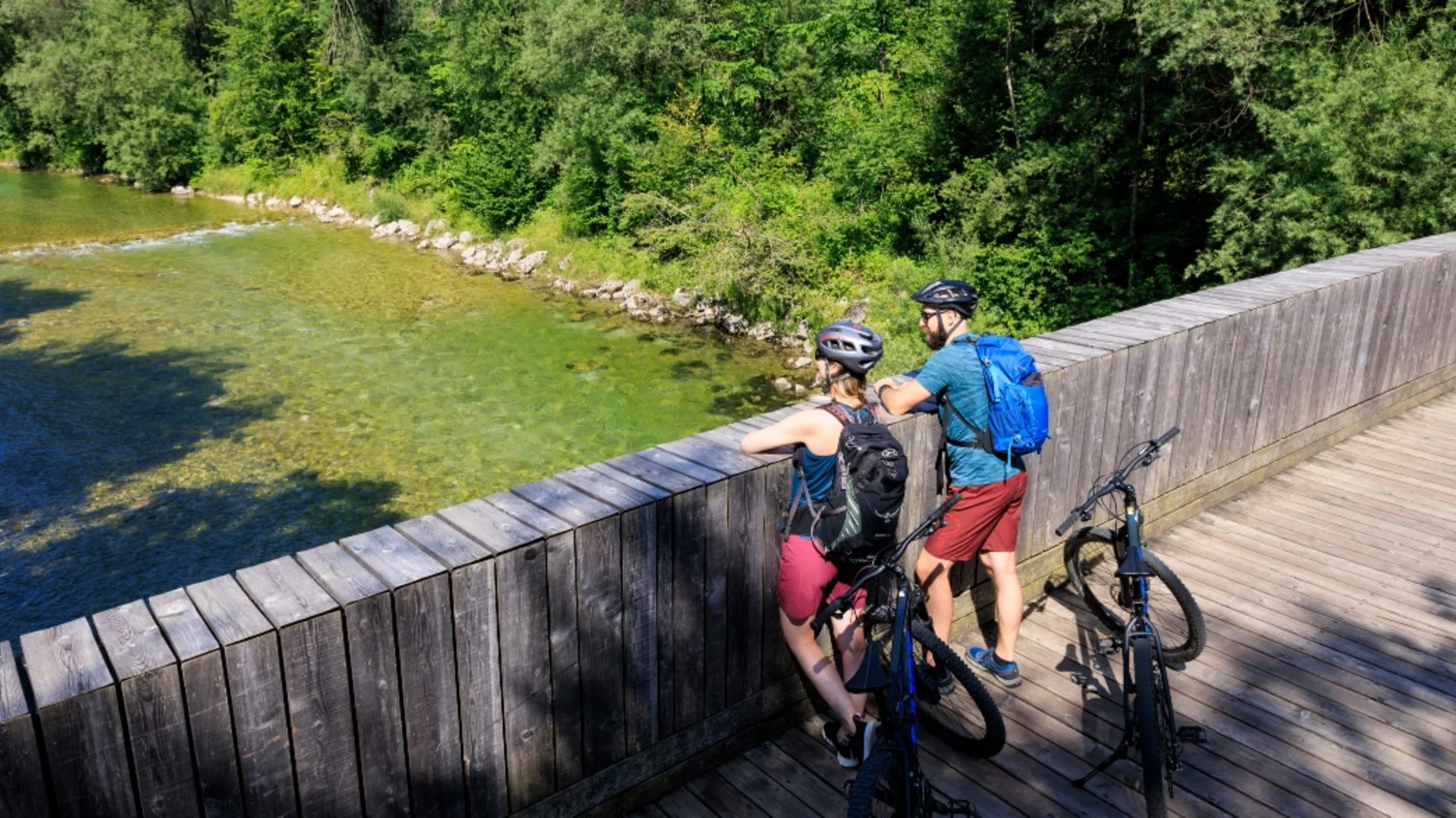Twee fietsers met helmen en rugzakken op een houten brug over helder water