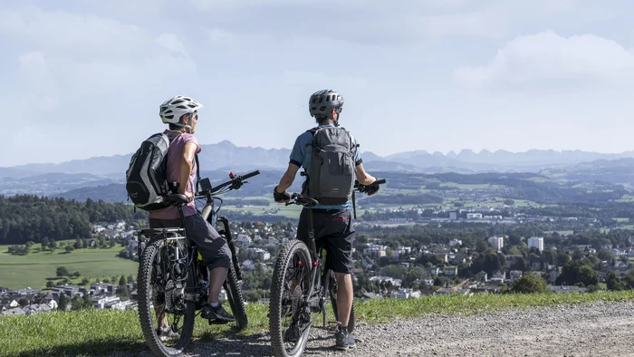 Zwei Radfahrer sehen von einem Hügel auf eine Stadt und Berge im Hintergrund
