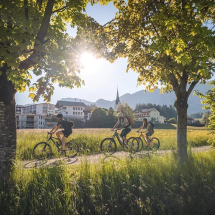 Drei Radfahrer fahren auf einem Weg durch eine grüne Wiese mit Bäumen und Dorf im Hintergrund