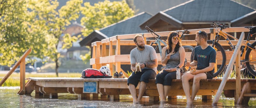 Three people sitting on dock with feet in water, bikes nearby, enjoying healthy snacks
