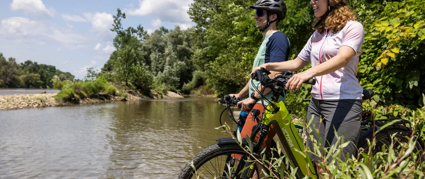Genuss mit jedem Tritt im Thermen- & Vulkanland © Heiko Mandl Zwei Radfahrer mit Helmen stehen mit ihren Fahrrädern an einem Fluss