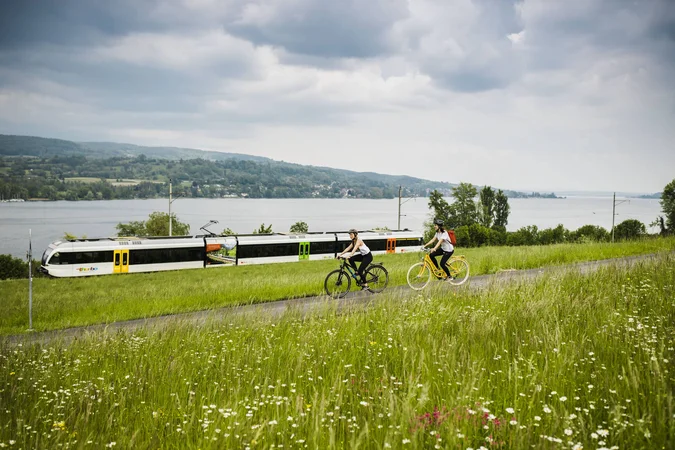Zwei Radfahrer fahren auf einem Weg neben einer Bahnstrecke am See