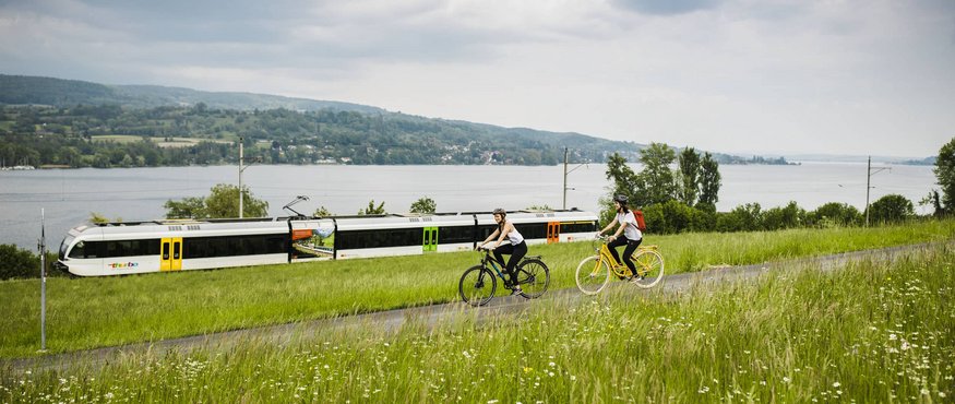 Two cyclists ride on a path beside a train line by the lake