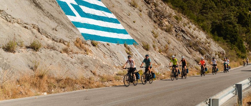 Cyclists on road beside cliff with large Greek flag painted