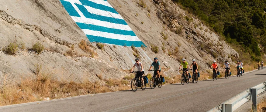 Fahrradfahrer auf Straße neben Felswand mit griechischer Flagge