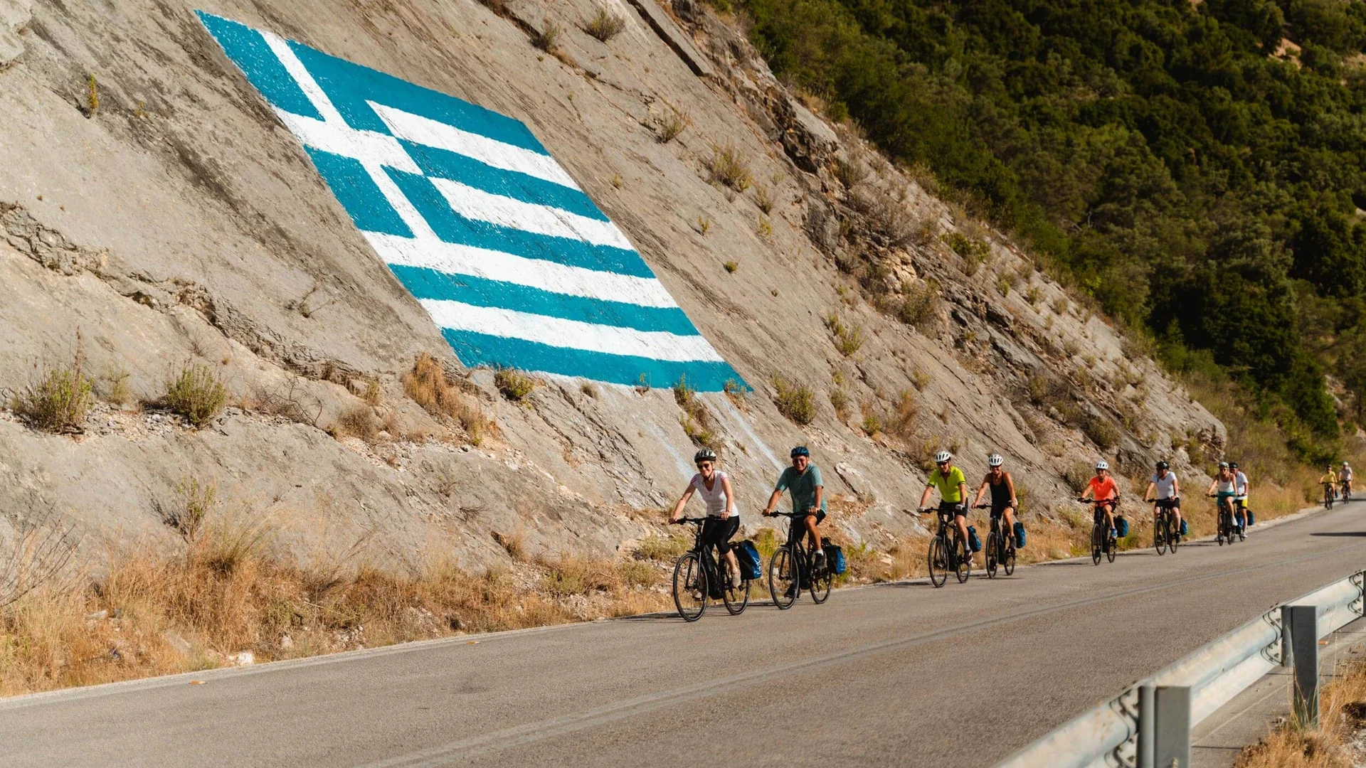 Fahrradfahrer auf Straße neben Felswand mit griechischer Flagge