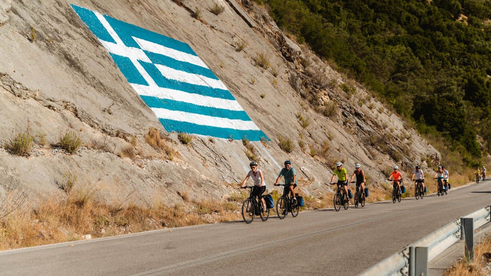 Cyclists on road beside cliff with large Greek flag painted
