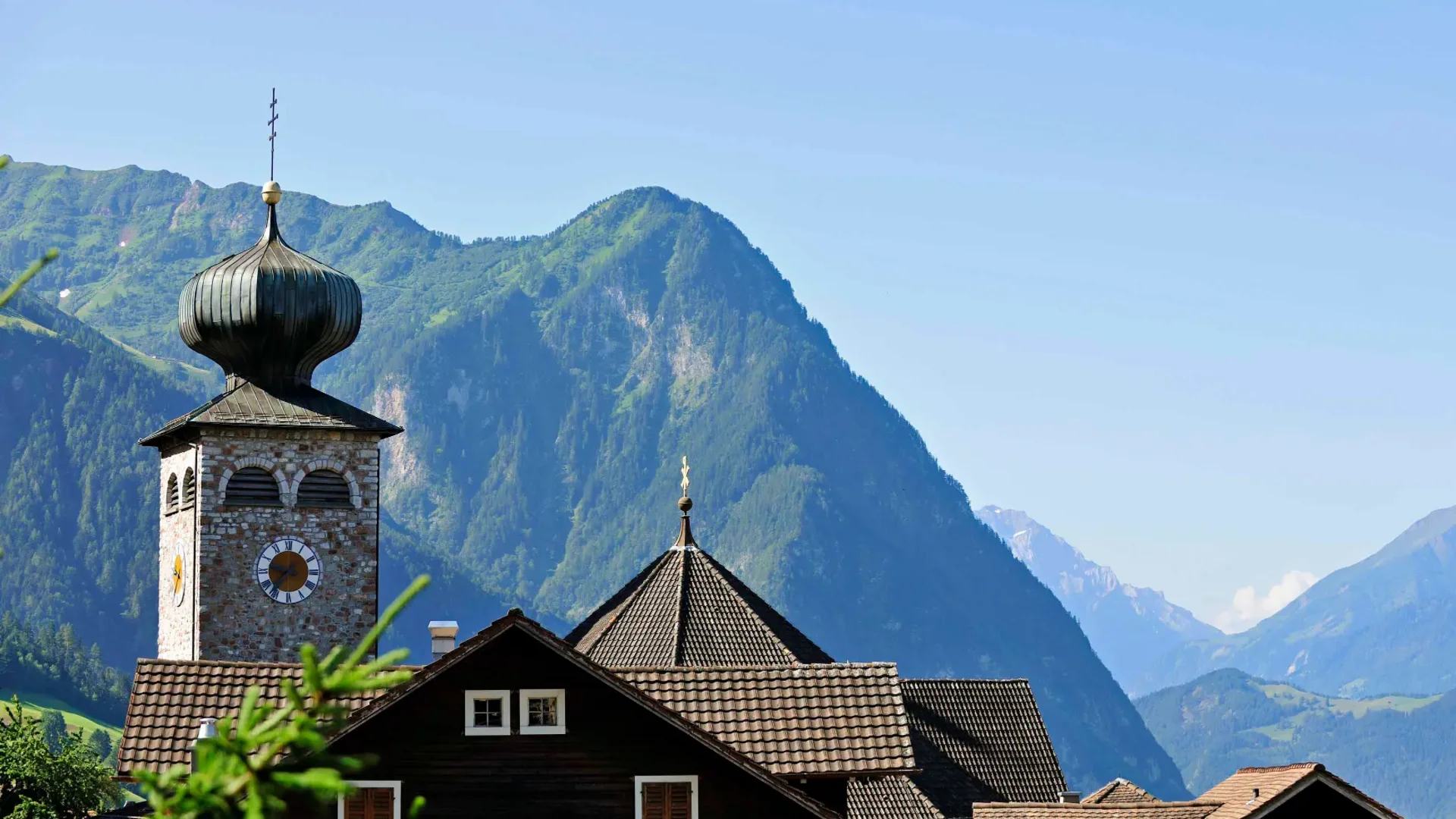 Kirchturm und Dächer vor alpiner Berglandschaft bei klarem Himmel