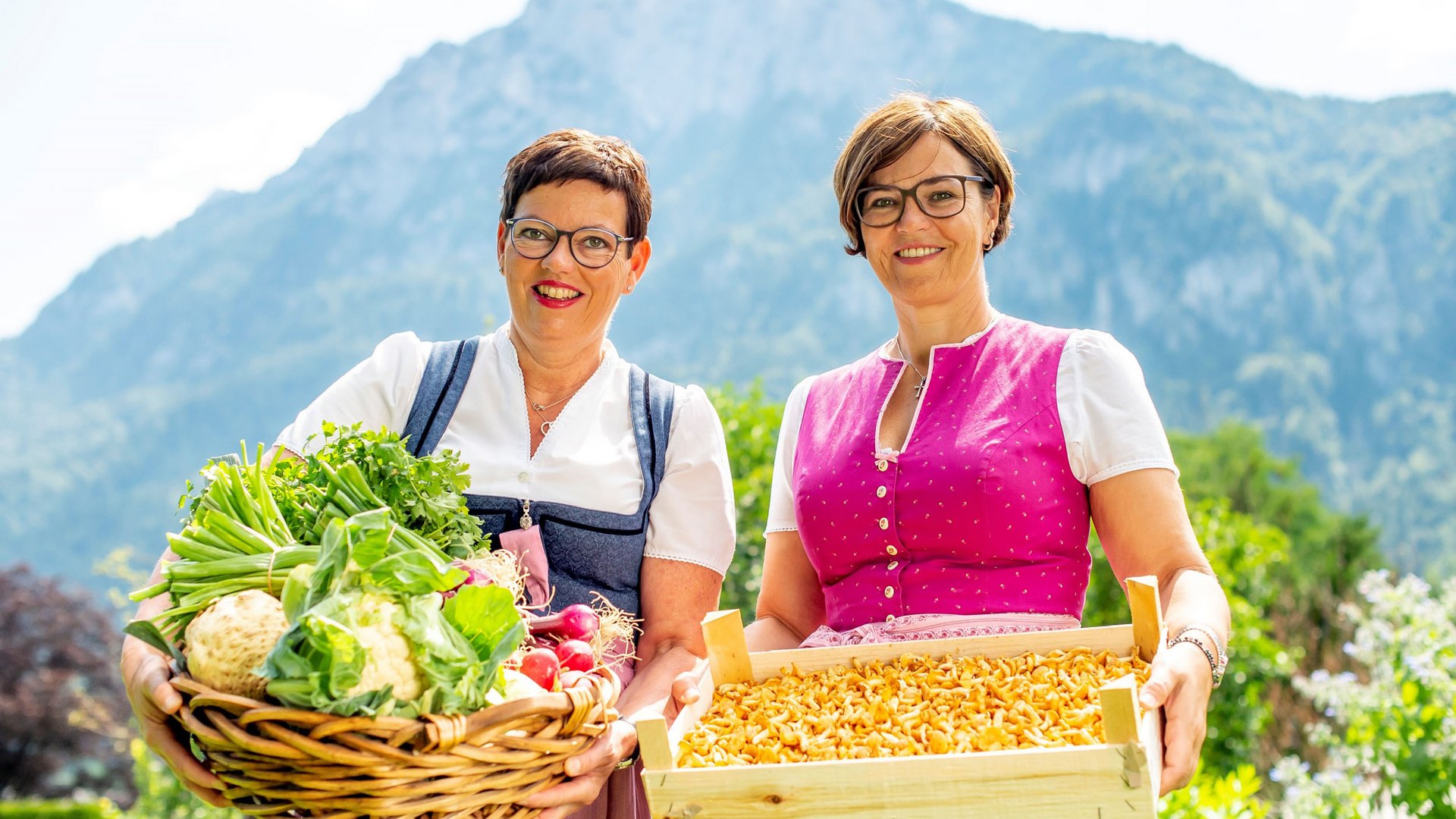 Zwei Frauen in Trachten halten frisches Gemüse und Pilze vor Bergkulisse