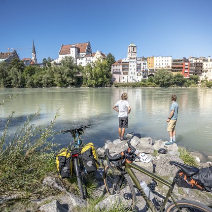 Zwei Radfahrer stehen am Fluss mit Blick auf eine Stadt am Ufer bei klarem Himmel