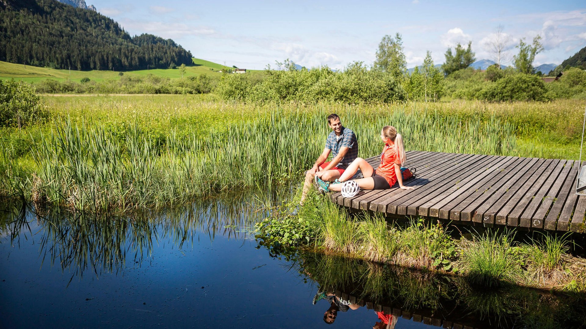 Two people sitting on a wooden dock by a lake in nature