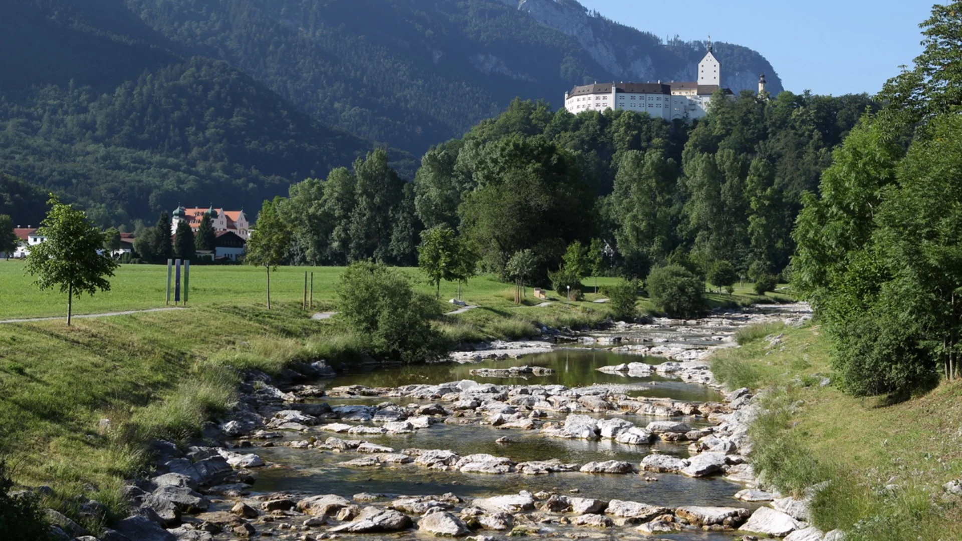 Fluss mit Felsen und Schloss auf bewaldetem Hügel vor Bergen