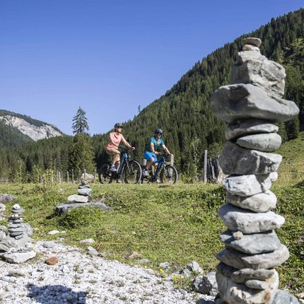 Zwei Radfahrer fahren in einem grünen Bergtal mit Steinmännchen im Vordergrund