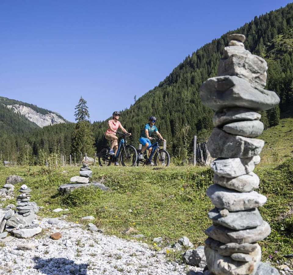 Zwei Radfahrer fahren in einem grünen Bergtal mit Steinmännchen im Vordergrund