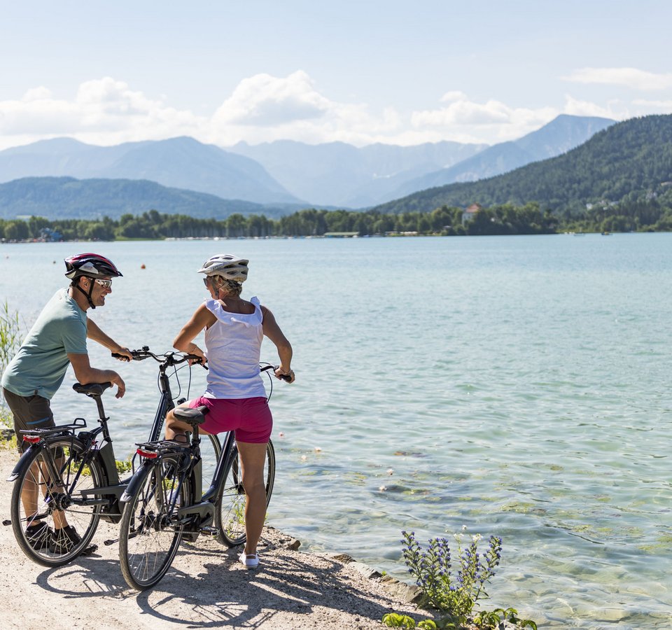 Zwei Radfahrer mit Helmen am Seeufer mit Bergen im Hintergrund