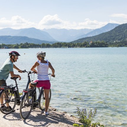 Zwei Radfahrer mit Helmen am Seeufer mit Bergen im Hintergrund