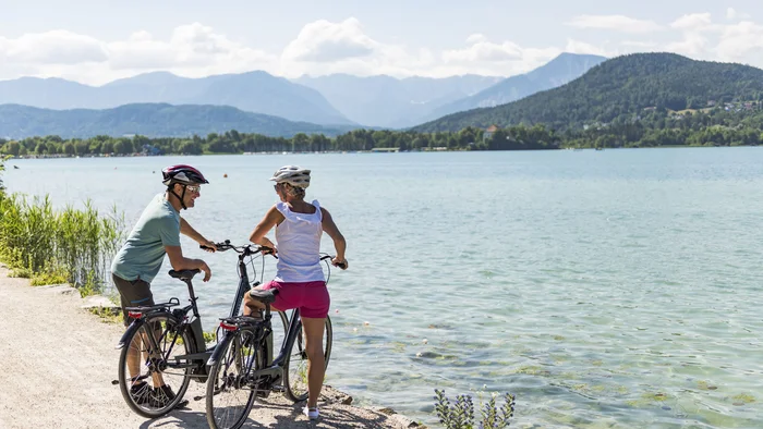 Klagenfurt kann mehr als Wörthersee! © Franz Gerdl Zwei Radfahrer mit Helmen am Seeufer mit Bergen im Hintergrund