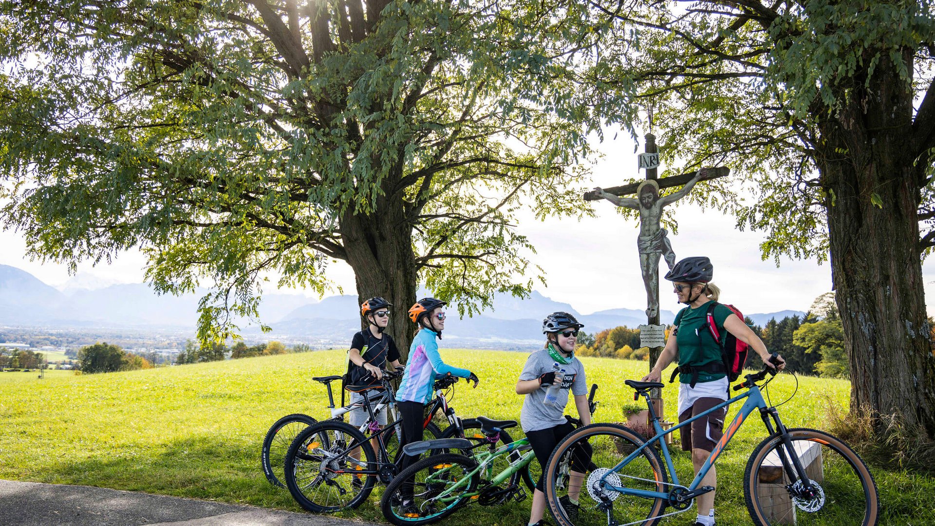Familie mit Fahrrädern macht Pause unter Bäumen neben einem Kreuz