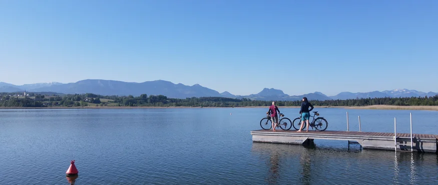 Zwei Personen mit Fahrrädern auf einem Steg am See mit Bergen im Hintergrund