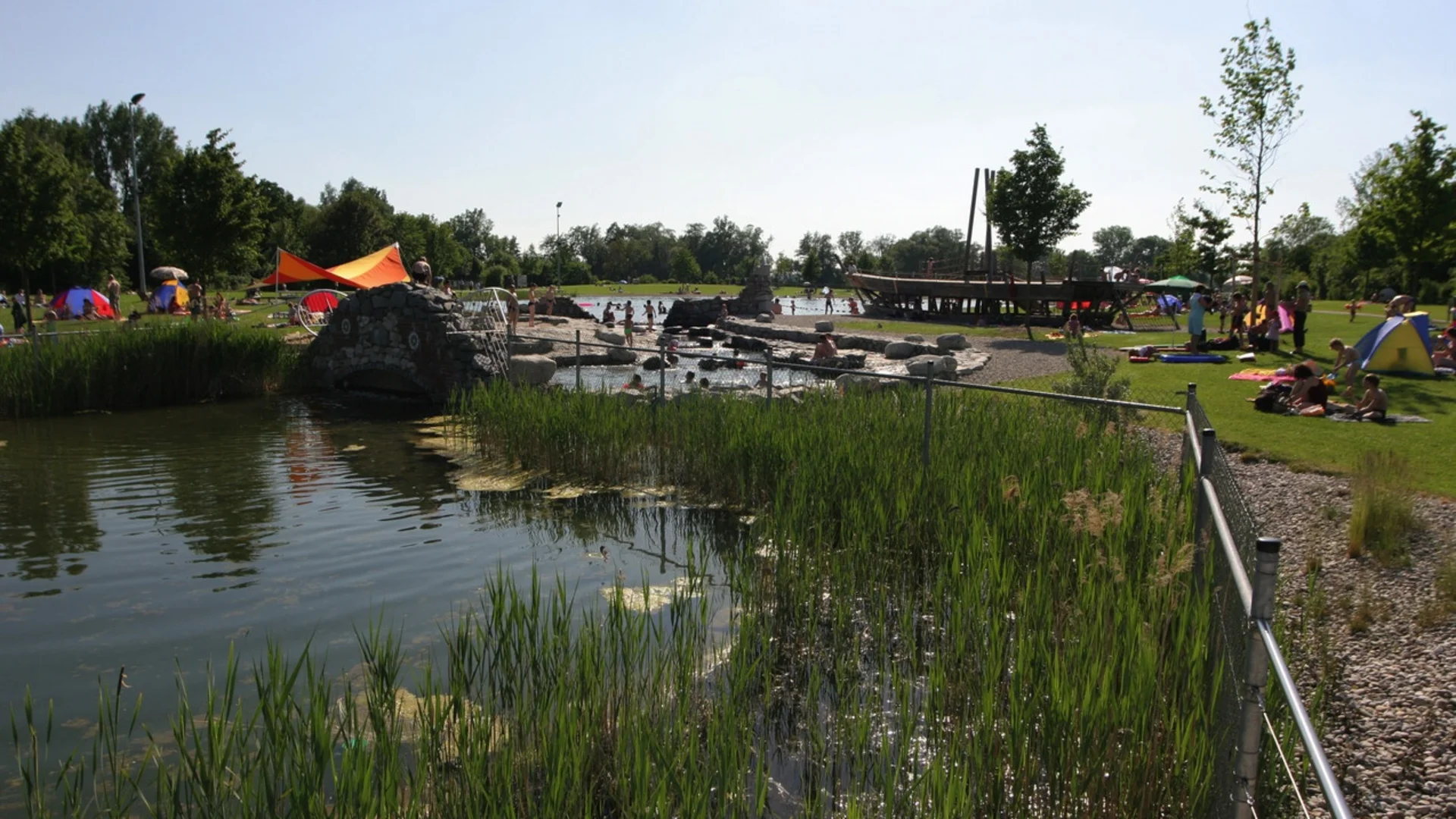 Grüner Park mit Teich, Kinder spielen im Wasser und Menschen entspannen auf der Wiese