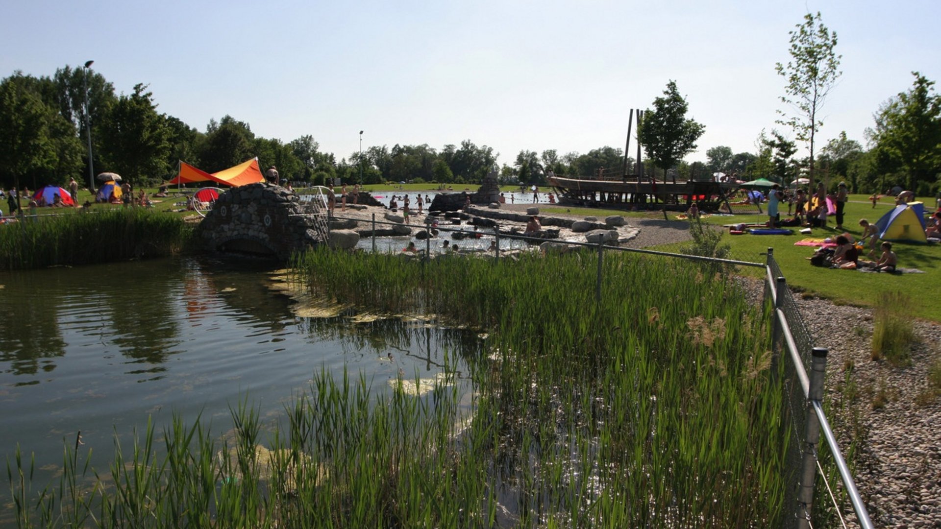 Green park with pond, children playing in water, and people relaxing on grass