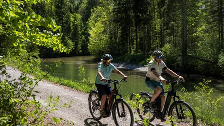Zwei Radfahrer fahren auf einem Waldweg neben einem Fluss an einem sonnigen Tag