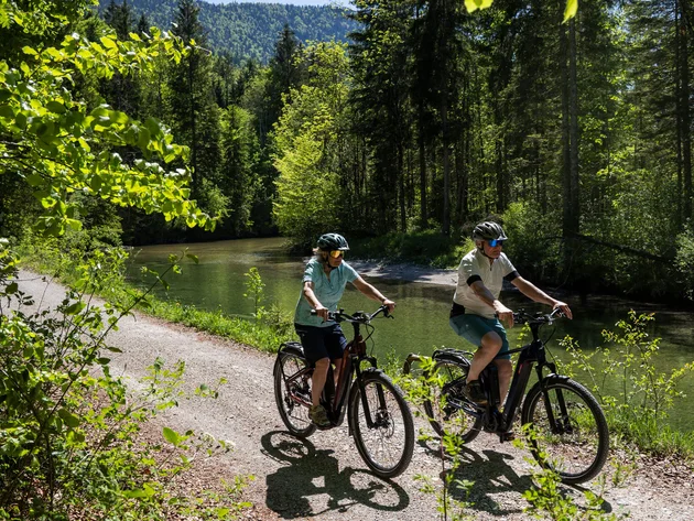 Zwei Radfahrer fahren auf einem Waldweg neben einem Fluss an einem sonnigen Tag