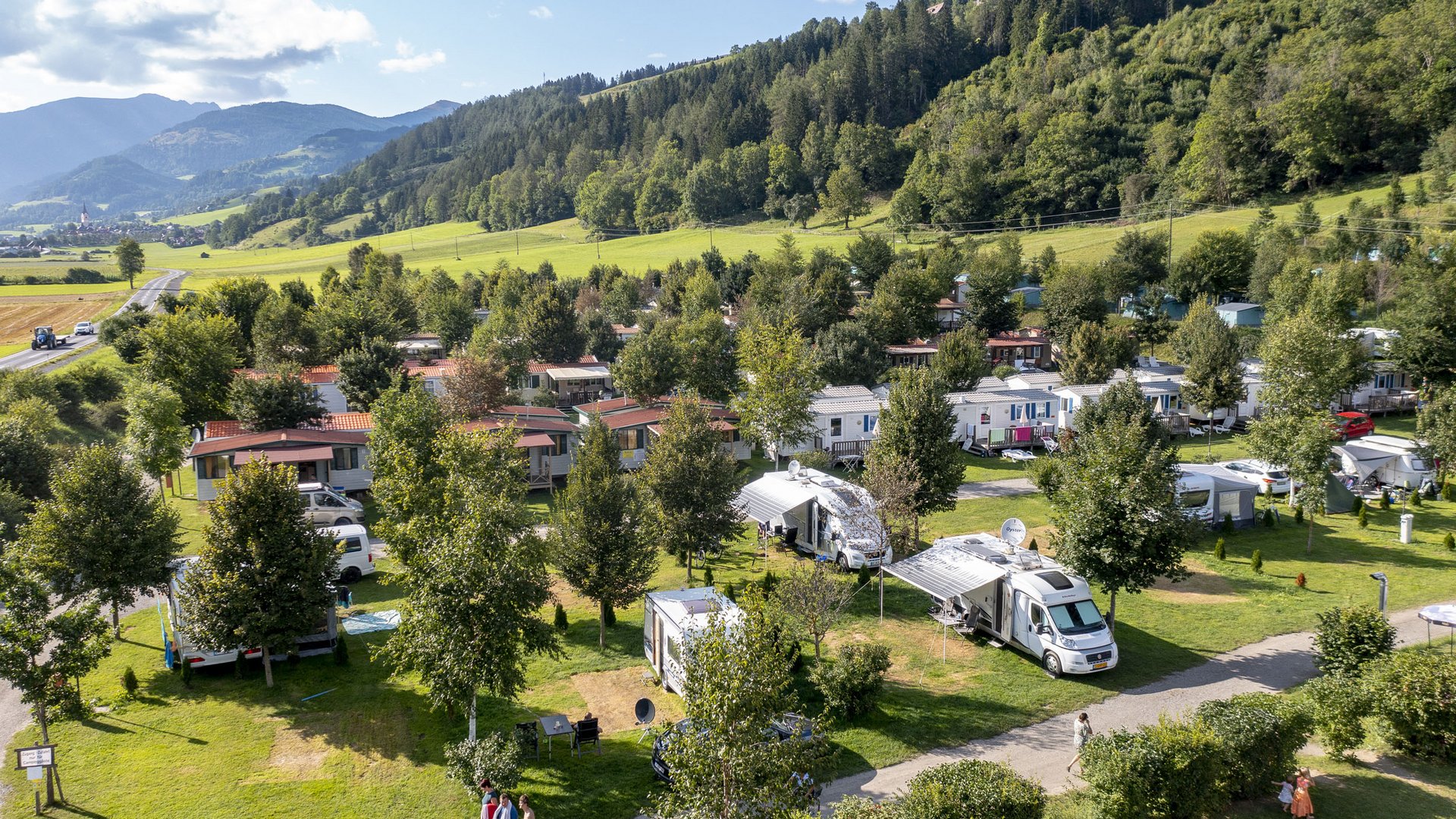 Campsite with motorhomes and trees near mountains on a sunny day