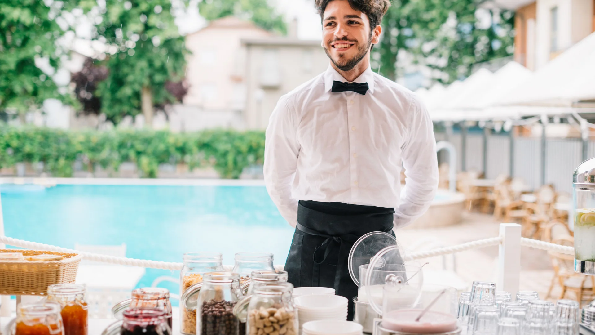 Young waiter standing at buffet with jars and glasses by pool
