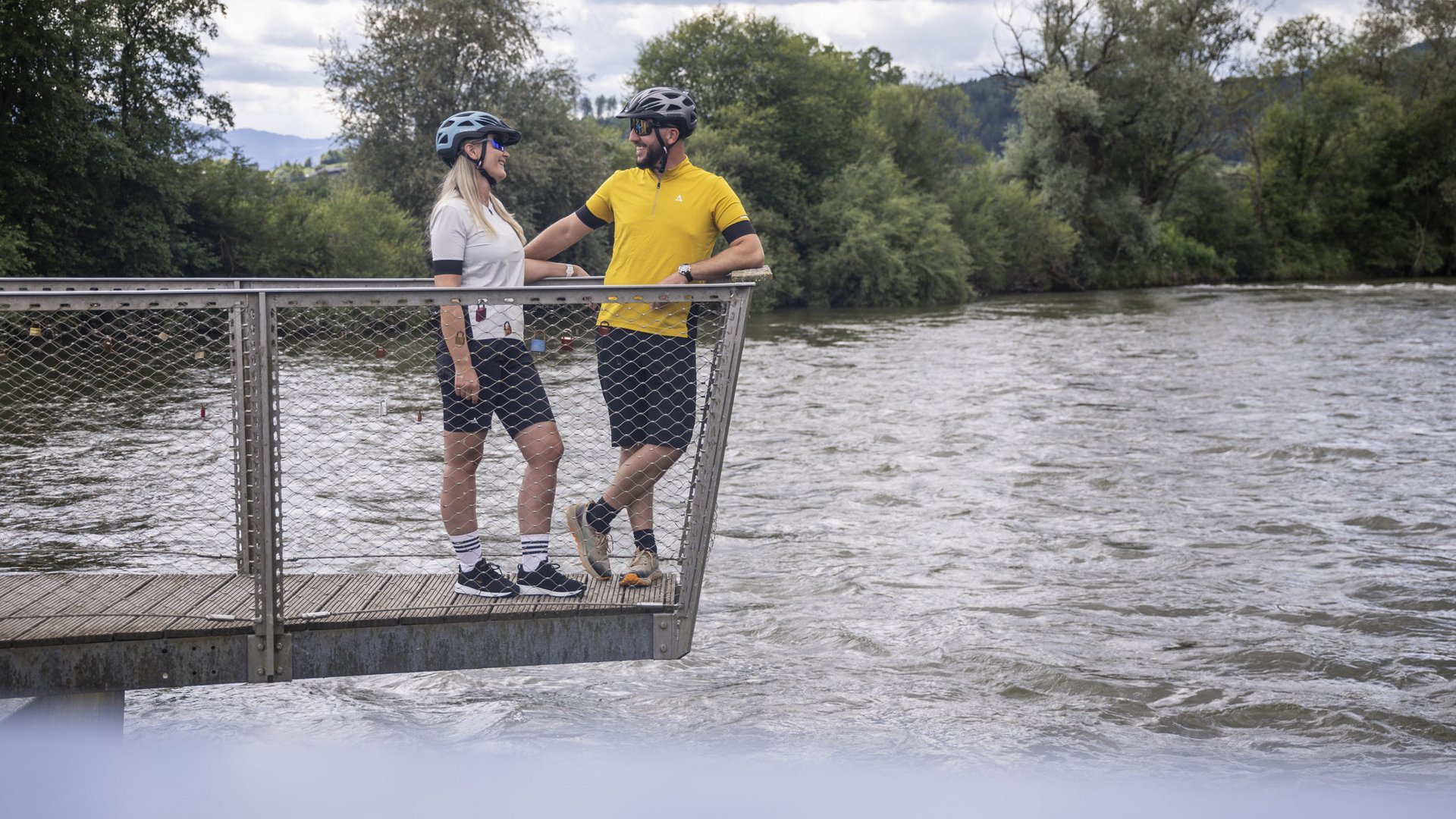 Two cyclists with helmets standing on a bridge over a river
