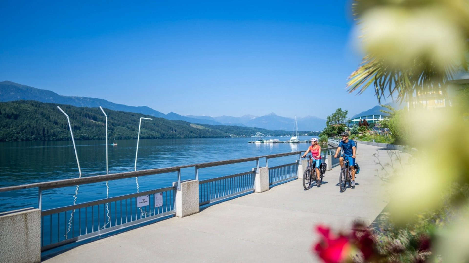 Two cyclists riding on a lakeside path with mountains and clear sky