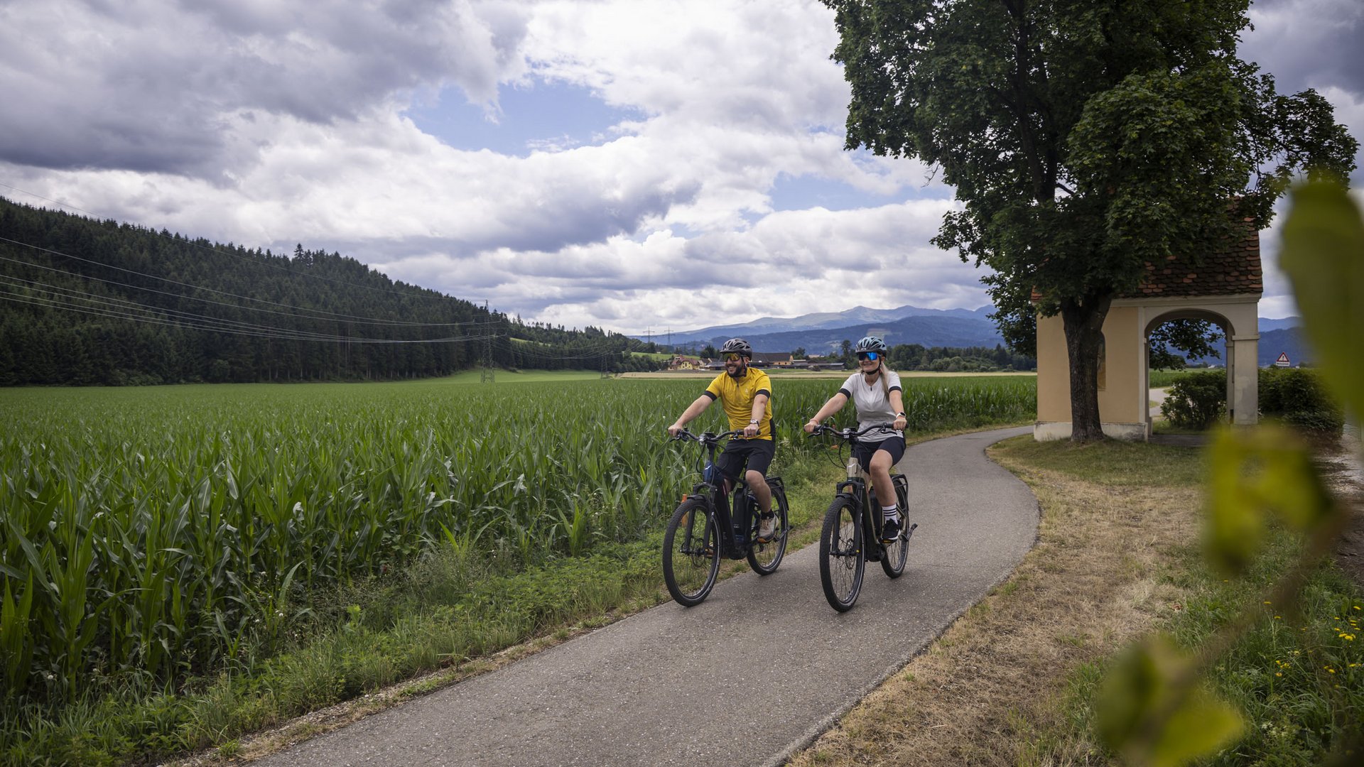 Two cyclists riding on a path through fields with mountains in the background