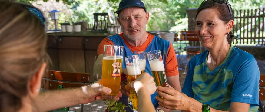 Drei Personen stoßen mit Biergläsern im Freien unter Bäumen an