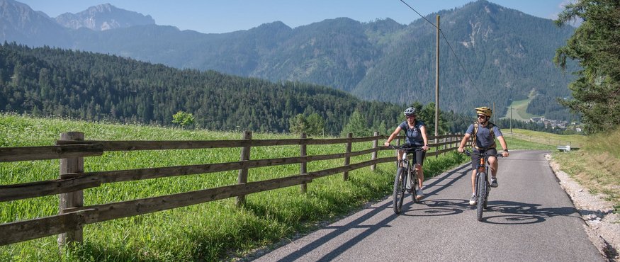 Two cyclists riding on mountain road with wooden fence and forest background