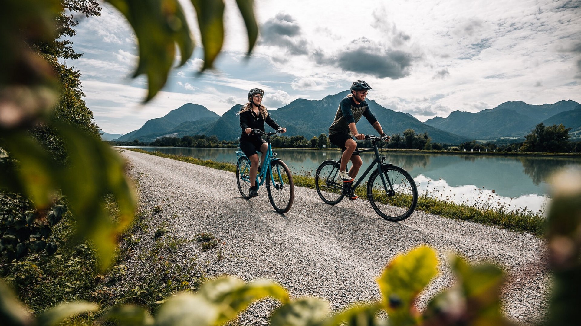 Two cyclists riding on a path beside a lake with mountains in the background
