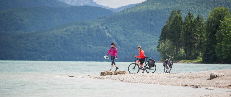 Two cyclists by the lakeshore with forested mountains in background