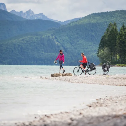 Zwei Radfahrer am Seeufer vor bewaldeten Bergen