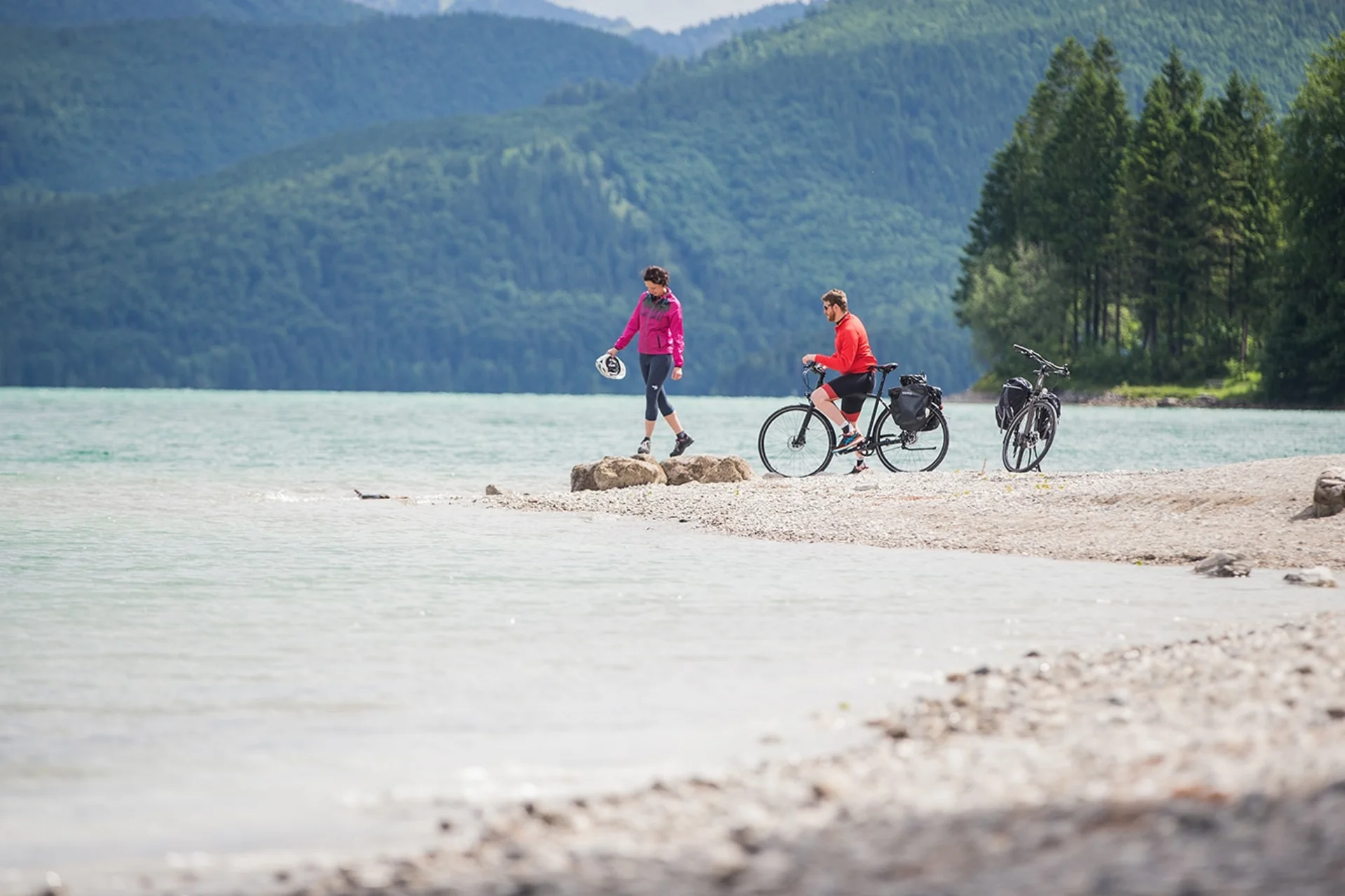 Zwei Radfahrer am Seeufer vor bewaldeten Bergen