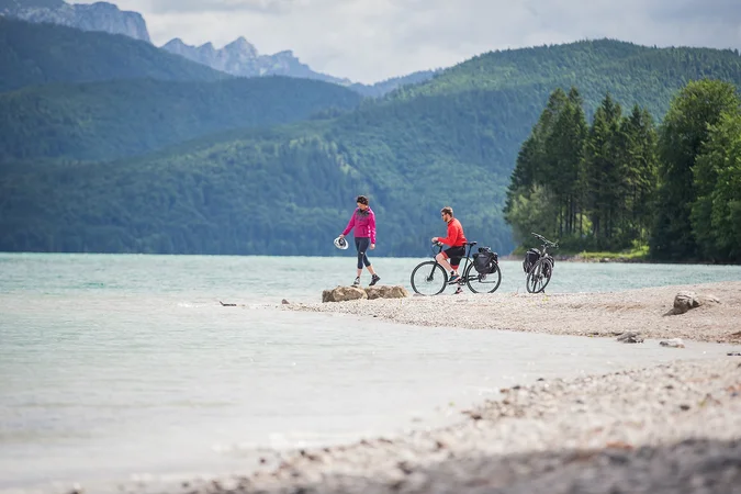 Zwei Radfahrer am Seeufer vor bewaldeten Bergen
